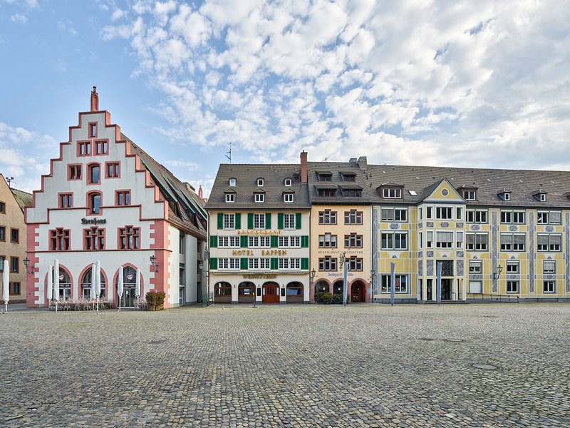 Freiburg: Der Münsterplatz mit dem 1970 wieder aufgebauten Kornhaus und historischer traufständiger Häuserzeile – sonst belebt durch Touristen, Wochenmarkt, Cafés und Restaurants