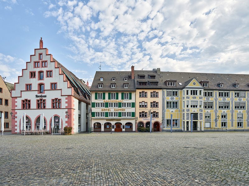 Freiburg: Der Münsterplatz mit dem 1970 wieder aufgebauten Kornhaus und historischer traufständiger Häuserzeile – sonst belebt durch Touristen, Wochenmarkt, Cafés und Restaurants